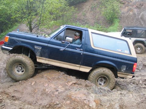 1991 Ford Bronco - A blue and tan Ford Bronco is shown off-roading on a challenging, muddy terrain, with another vehicle visible in the background.