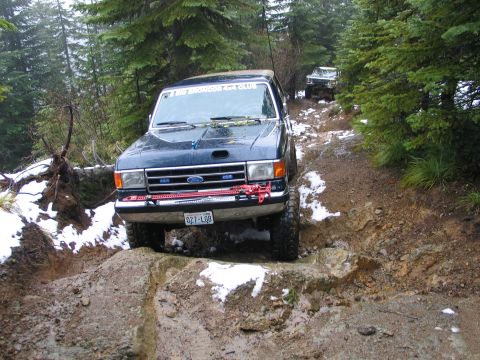 1991 Ford Bronco - Two Ford Broncos navigate a challenging off-road trail in a wooded area during winter.