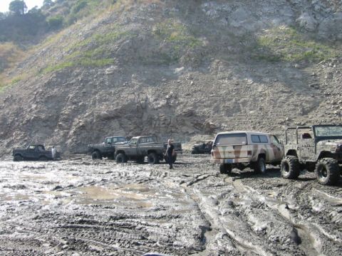 1985 Ford Bronco - Several off-road vehicles are stuck in thick mud near a rocky hillside.