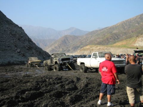 1985 Ford Bronco - Multiple trucks are bogged down in thick mud, with a line of other vehicles visible in the distance.