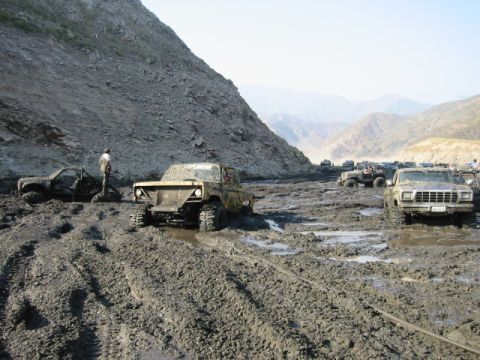 1985 Ford Bronco - Several off-road vehicles navigate a treacherous muddy terrain, with one 1980s pickup truck prominently stuck in the mire.