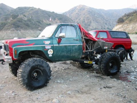 1985 Ford Bronco - Two off-road vehicles are parked on a rocky surface near hills.