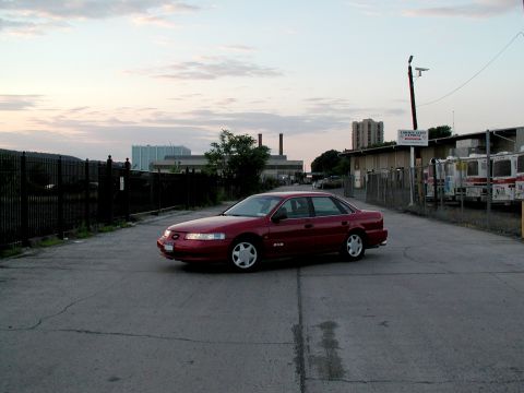 1992 Ford Taurus - Red first-generation Ford Taurus SHO sedan parked at an angle on cracked concrete in an industrial lot at dusk, wearing silver five-spoke S…