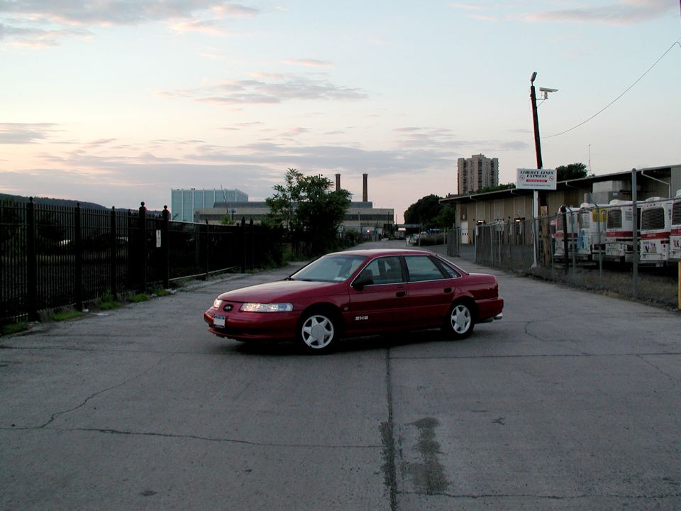 Red Ford Taurus SHO sedan parked in industrial lot at dusk, front three-quarter view