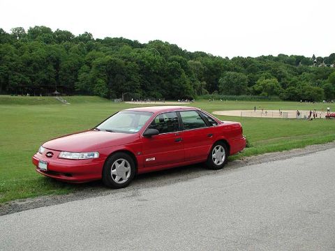 1992 Ford Taurus - Red 1992 Ford Taurus parked beside a road with a baseball game in progress on the field behind it.