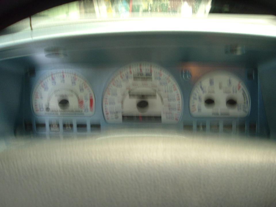 Car dashboard with three main gauges, including speedometer and tachometer. Some text is visible on the gauges