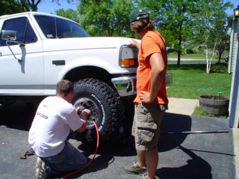 1993 Ford Bronco - A man inflates a large tire on a white Ford F-Series truck, while another man stands nearby.