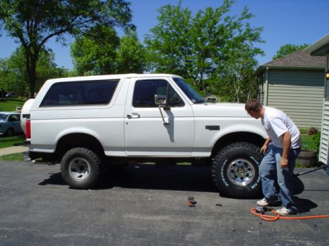1993 Ford Bronco - A man inspects the large tires on a lifted white 1993 Ford Bronco parked in a driveway.