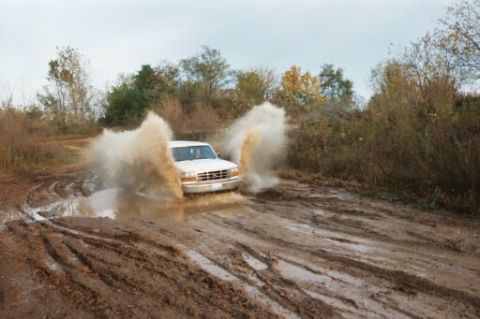 1993 Ford Bronco - A white Ford F-Series truck navigates a muddy track, kicking up a large spray of water and mud.