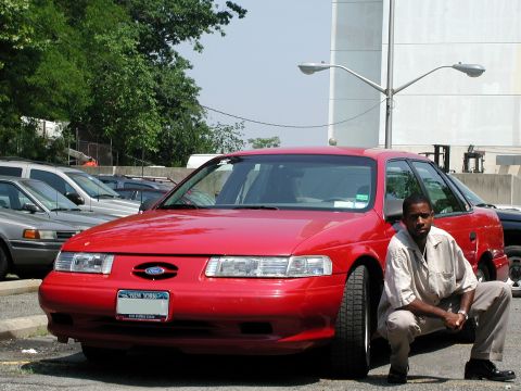 1992 Ford Taurus - A red 1992 Ford Taurus sedan is parked in a lot, with a man posing next to it.