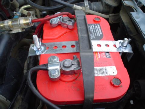 1993 Ford Bronco - Close-up of a car battery with wiring and hold-down bracket.