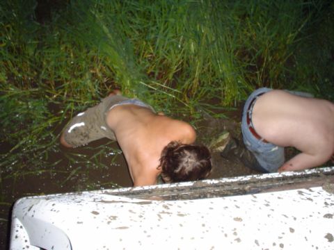 1993 Ford Bronco - Two individuals are partially submerged in mud and grass, with a portion of a white vehicle visible in the foreground.