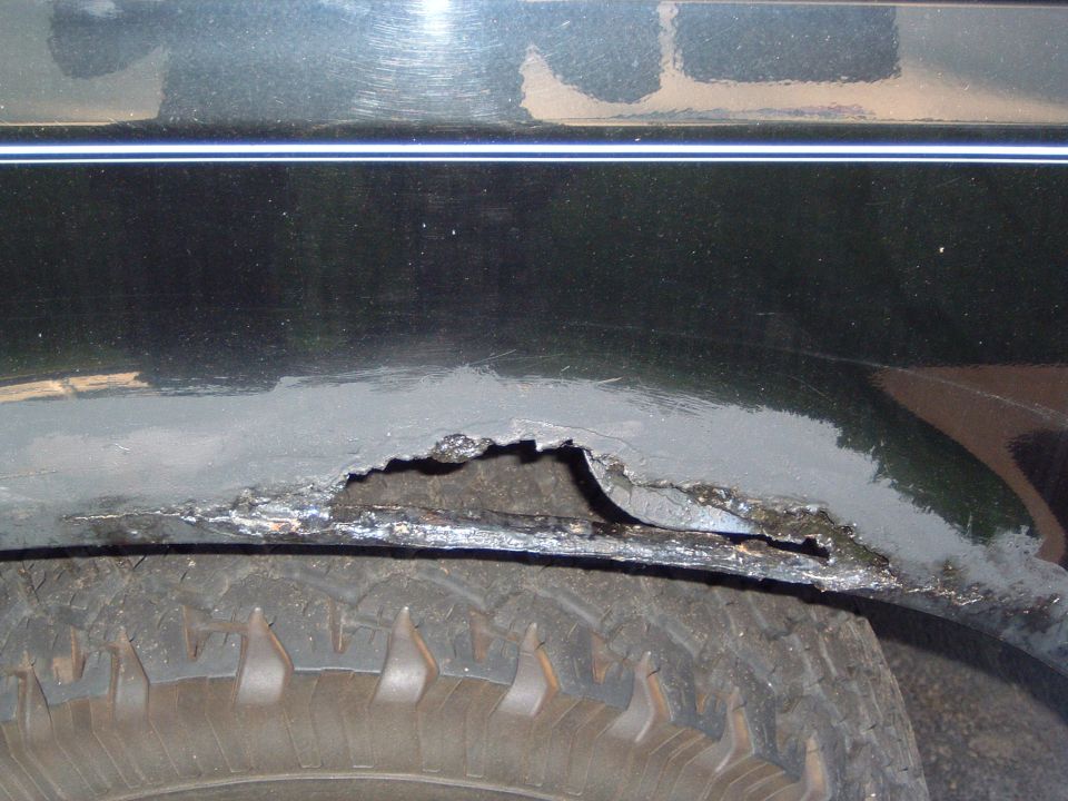 Severe rust holes and body filler on dark green fender lip above an off-road tire