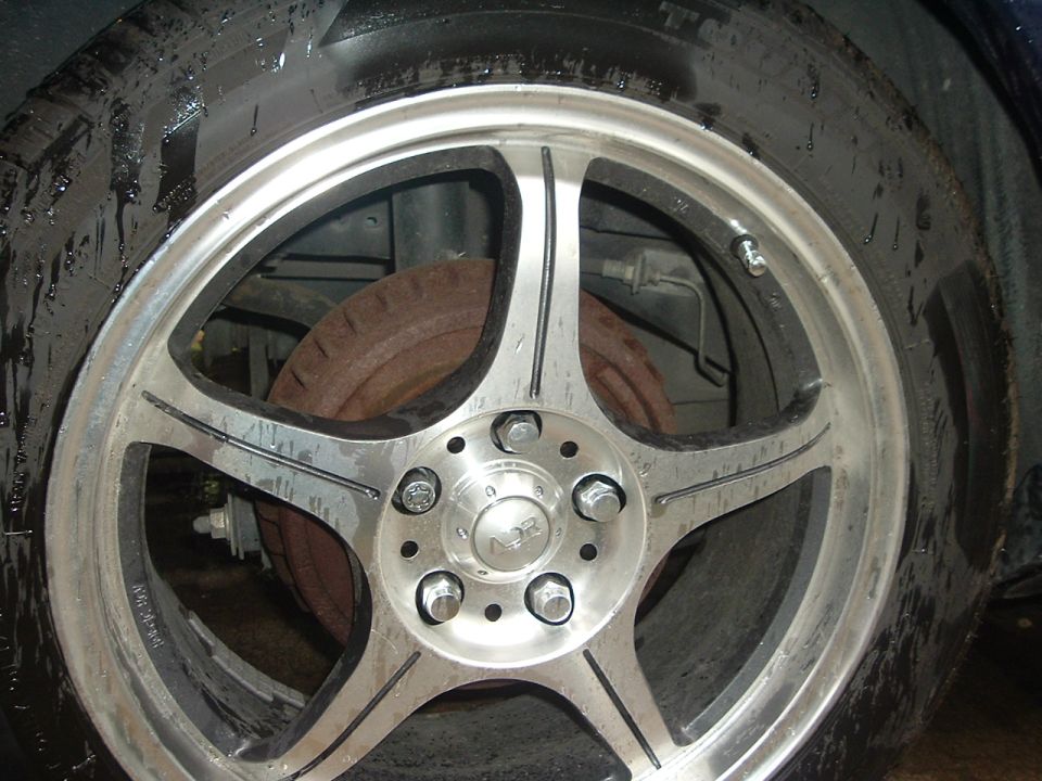 Close-up of a silver five-spoke aftermarket wheel with a black tire. The wheel has the letters 'NSR' in the center cap. Rusty brake rotor visible behind the wheel