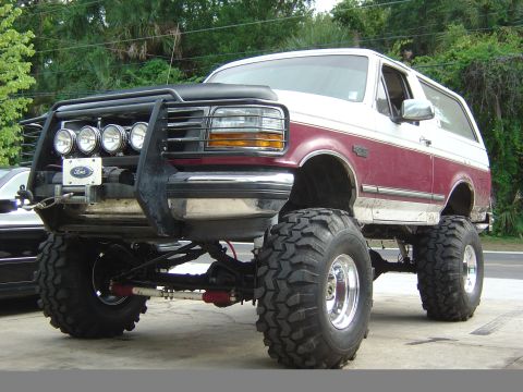 1993 Ford Bronco - A lifted two-tone red and white 1990s Ford Bronco SUV shown from a 3/4 front angle, riding on large mud-terrain tires with polished wheels,…