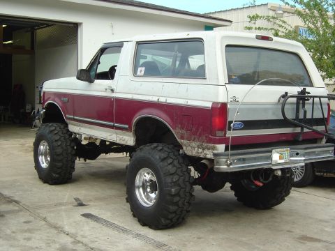 1993 Ford Bronco - Three-quarter rear view of a lifted two-tone burgundy and white full-size Bronco on a concrete shop apron, showing the removable hardtop, t…