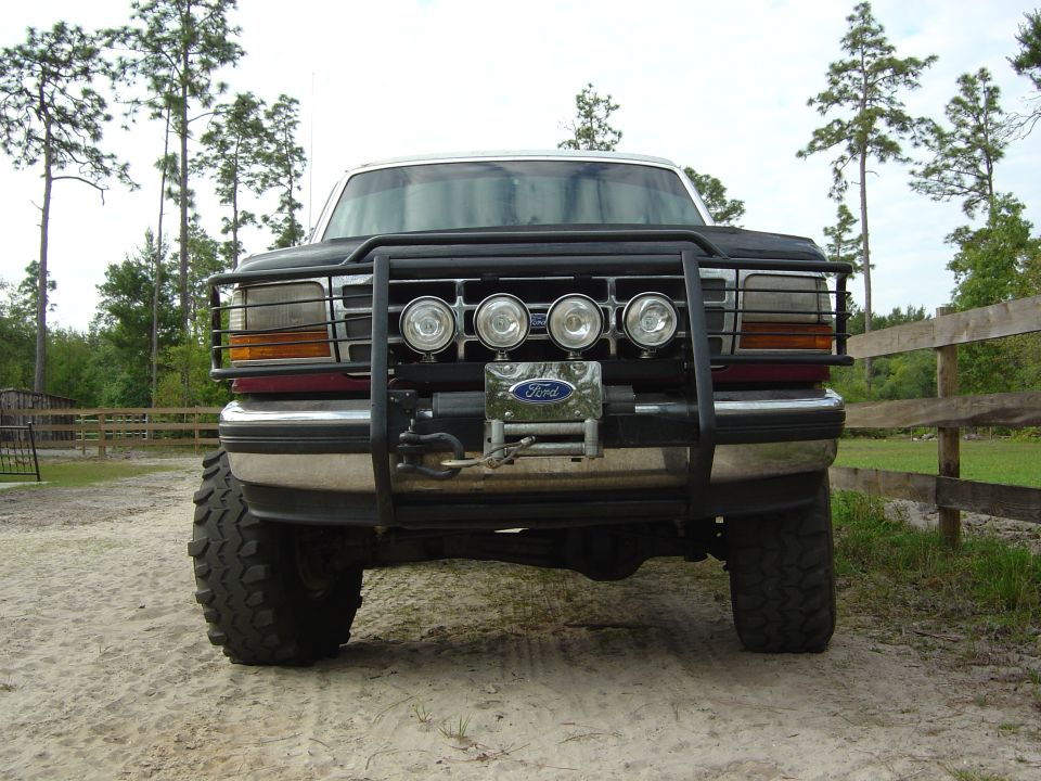 Front view of a dark colored Ford truck with a black bull bar, four auxiliary lights, and a winch. It has large off-road tires