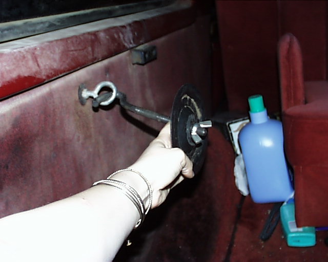 Hand holding a black circular part with a metal rod attached to a hook on a red interior panel. Blue bottle and green stapler visible