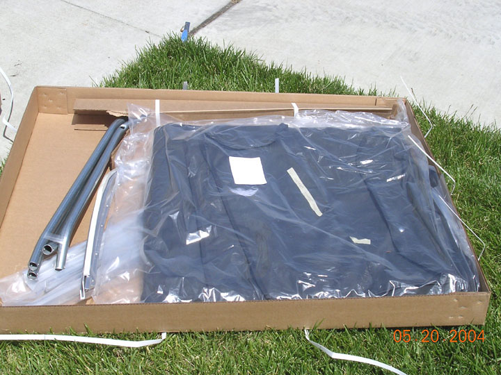 Gray folded soft top in plastic wrap inside an open cardboard box on grass with metal bows