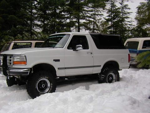 1995 Ford Bronco - A white 1995 Ford Bronco with a black soft top navigates through a snowy landscape, with another vehicle visible in the background.