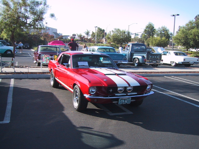 Red classic Mustang coupe with white racing stripes and grille fog lights at an outdoor car show