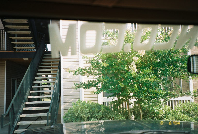Car interior view through windshield showing reflection of "MOD.COM" text and exterior scene with stairs and building