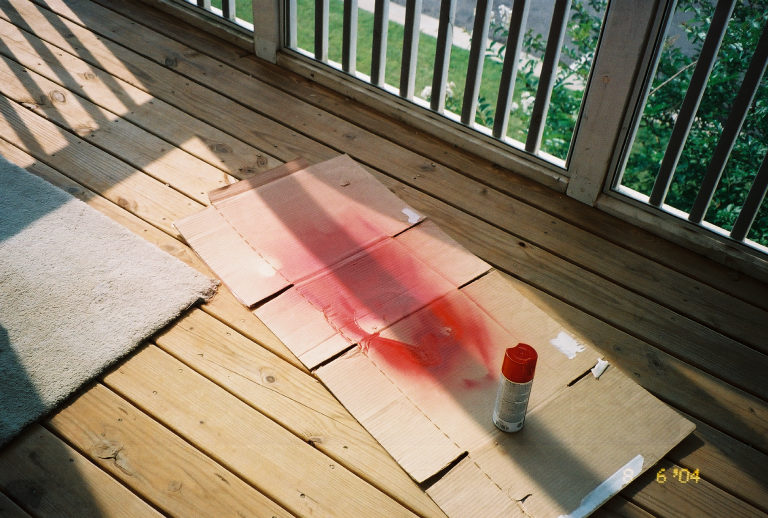 Red spray paint on cardboard with a can and brush on a wooden deck