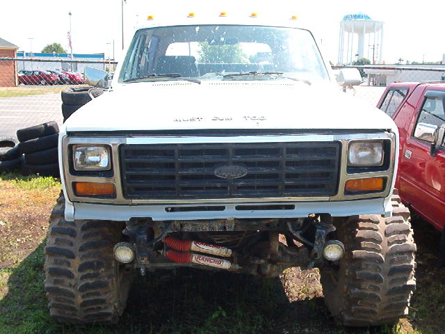 Front view of a lifted Ford F-Series truck with large off-road tires. A red vehicle is partially visible to the right