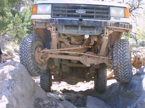 1987 Ford Bronco - A white 1987 Ford Bronco navigates rocky terrain, with its front suspension and grille clearly visible.