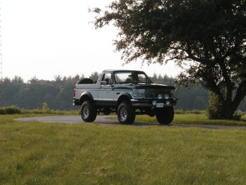 1994 Ford Bronco - This lifted Ford Bronco is parked on a road with a tree and grassy field in the background.