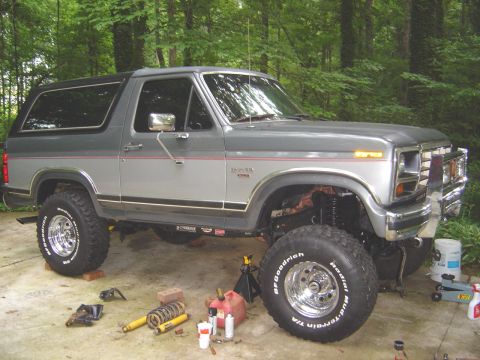1986 Ford Bronco - A two-tone gray and silver 1980s Ford Bronco full-size SUV with a lifted suspension and chrome-wheeled BFGoodrich Mud-Terrain T/A tires, sh…