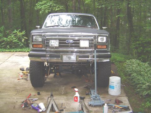 1986 Ford Bronco - Front view of a dark gray/black Ford SUV with a chrome grille and bumper, front end raised on a jack with tools, buckets, and a spray bottl…