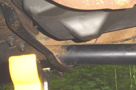 1986 Ford Bronco - Underside view of a vehicle showing a black driveshaft running horizontally, rusty frame components, bolts, and a fuel tank or skid plate a…