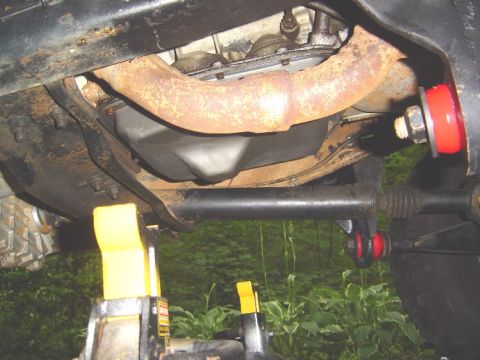 1986 Ford Bronco - Underside view of a vehicle showing a rusty exhaust/exhaust manifold pipe running across a cast differential or gearcase, with a black sway…