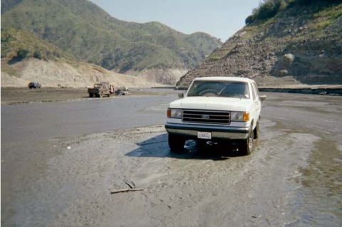 1990 Ford Bronco - A white Ford F-Series truck navigates a dry riverbed with mountainous terrain in the background.