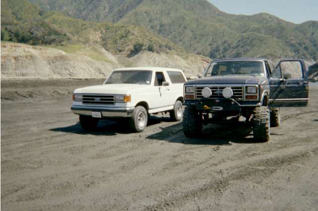 White and lifted dark blue full-size Ford Broncos parked side by side off-road