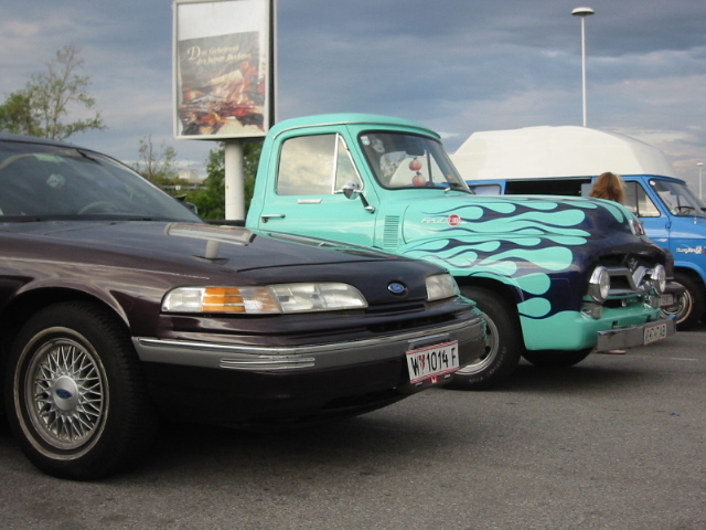 Dark sedan parked next to a teal pickup with blue flames. A blue van is behind the truck