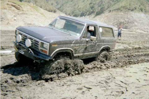 1985 Ford Bronco - A dark gray Ford Bronco is stuck deep in mud, with a person standing in the background.