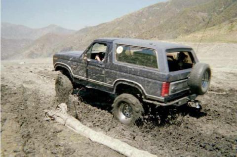 1985 Ford Bronco - A 1985 Ford Bronco navigates a challenging muddy terrain, with its rear spare tire prominently displayed.