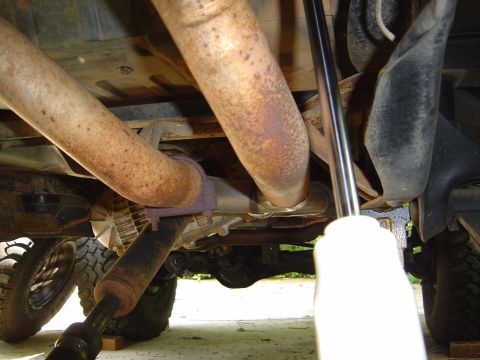 1986 Ford Bronco - Underside view of a vehicle showing rusty dual exhaust pipes routed over a rear axle, with a driveshaft, coil spring, and tire visible; a s…
