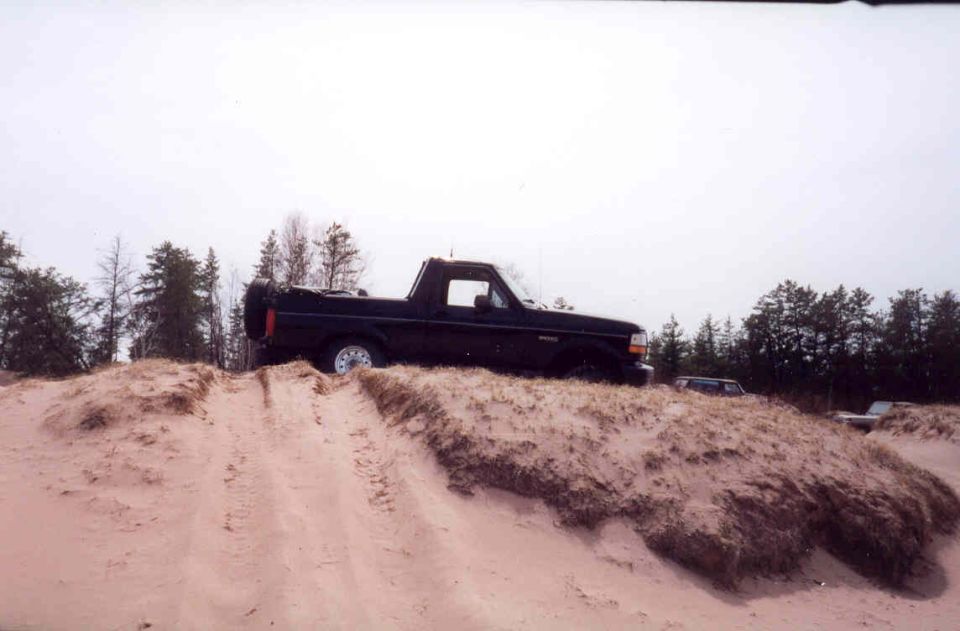 Black Ford Bronco on a sand dune, side view. Spare tire on rear. Other vehicles visible in background