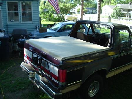 1991 Ford Bronco - A black Ford Bronco with a tan tonneau cover is parked in a yard next to a blue sedan.