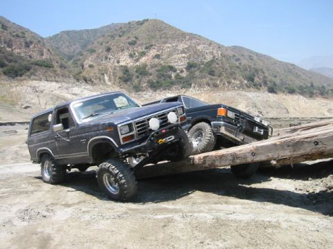 1985 Ford Bronco - Two SUVs tackle a challenging wooden obstacle on a dirt path with hills in the background.