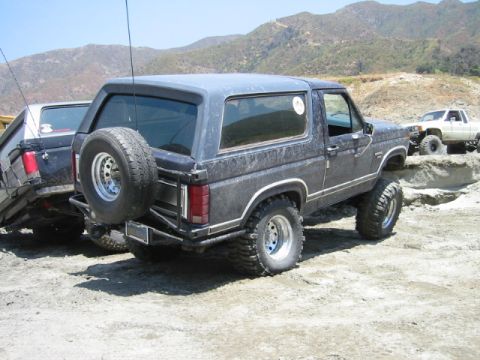 1985 Ford Bronco - Two off-road vehicles are parked on a dirt path with hills in the background.