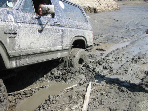 1985 Ford Bronco - A blue Ford Bronco is deeply stuck in mud, with its right rear tire submerged.