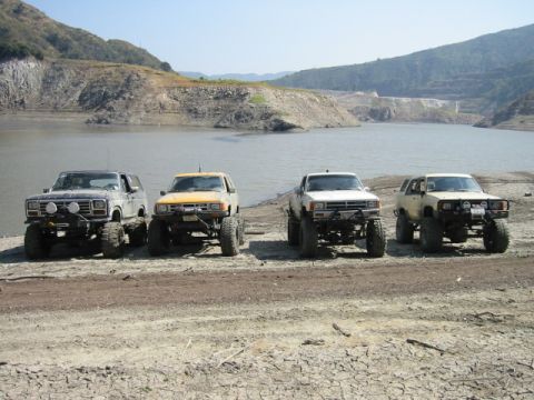 1985 Ford Bronco - Four modified SUVs are parked in a line near a lake with a dam in the background.