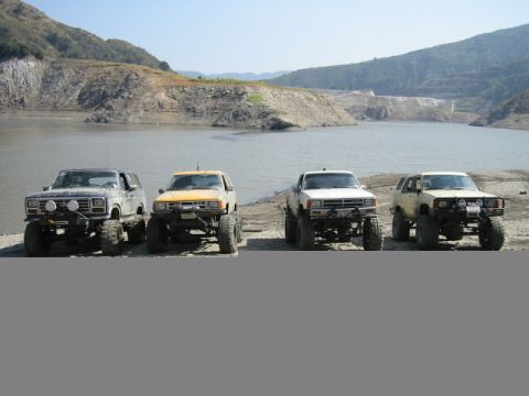1985 Ford Bronco - Several modified SUVs and trucks are parked on a dirt lot next to a large body of water under a clear sky.