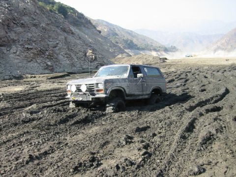 1985 Ford Bronco - Ford Bronco is stuck deep in mud with mountains in the background.