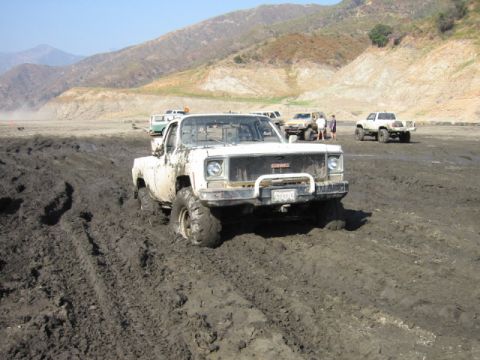 1985 Ford Bronco - A white GMC pickup truck is stuck in deep mud with other vehicles visible in the distance.