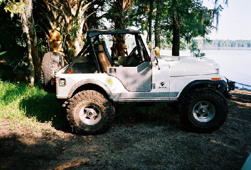 Silver Jeep CJ-5 with diamond-plate panels parked under trees by water